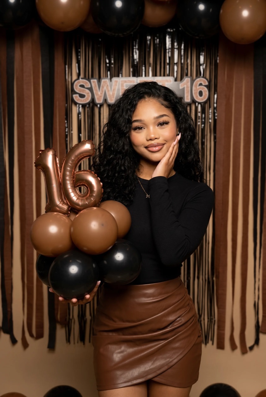 Teenage girl posing at a Sweet 16 birthday party with rose gold number balloons and elegant black and gold decorations.
